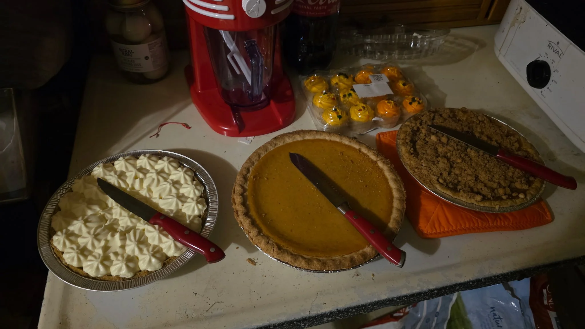 A variety of homemade pies and desserts on a kitchen counter, including pumpkin pie, cream pie, pecan pie, and cupcakes, with a red cupcake decorating tool in the background.