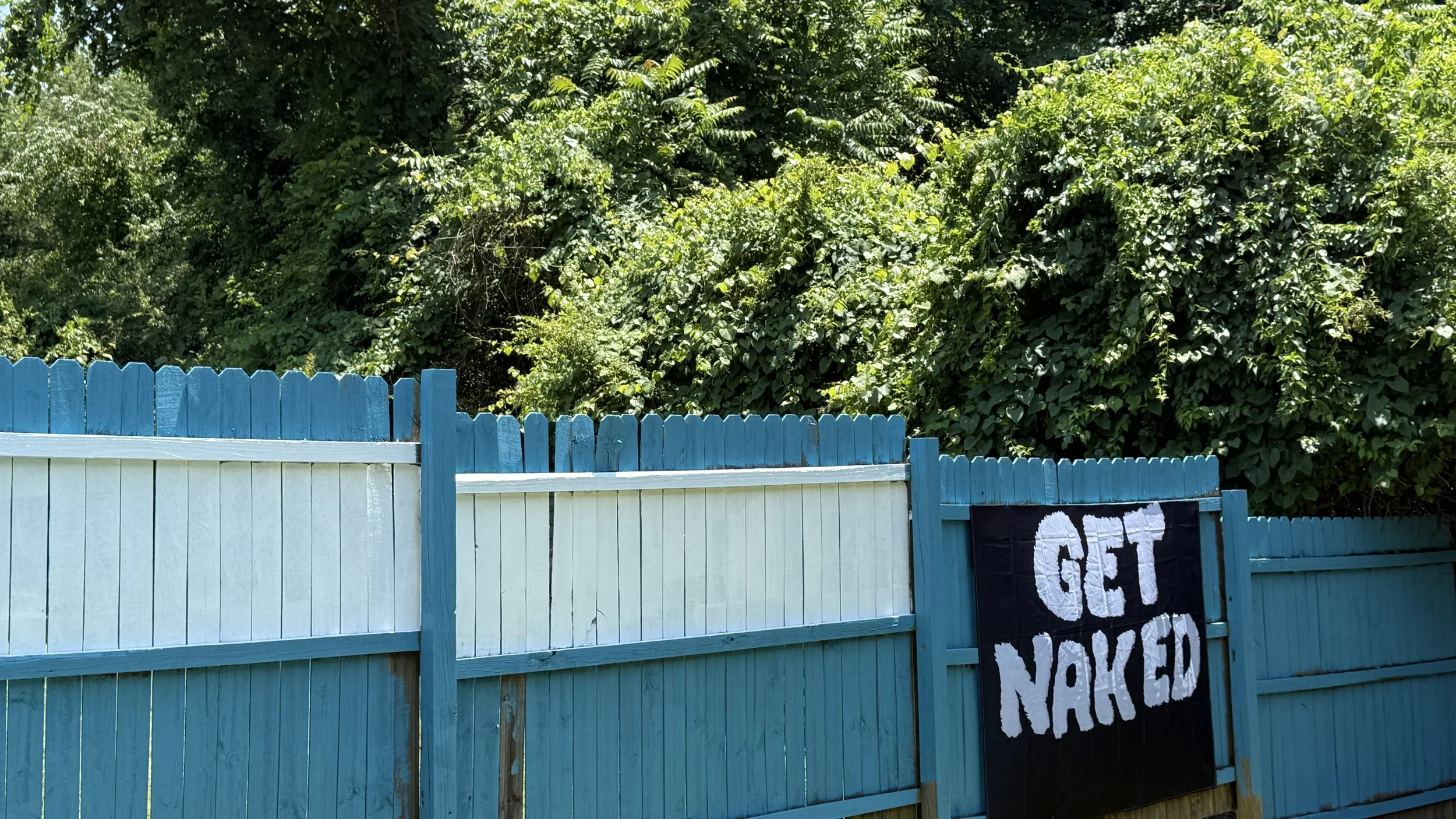 A white wooden fence with blue trim, partially covered by a black and white protest sign saying "GET NAKED". Behind the fence, there are trees and green foliage in the background.