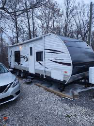 A white and black travel trailer parked on gravel with a gray car parked beside it, in a wooded area with leafless trees.