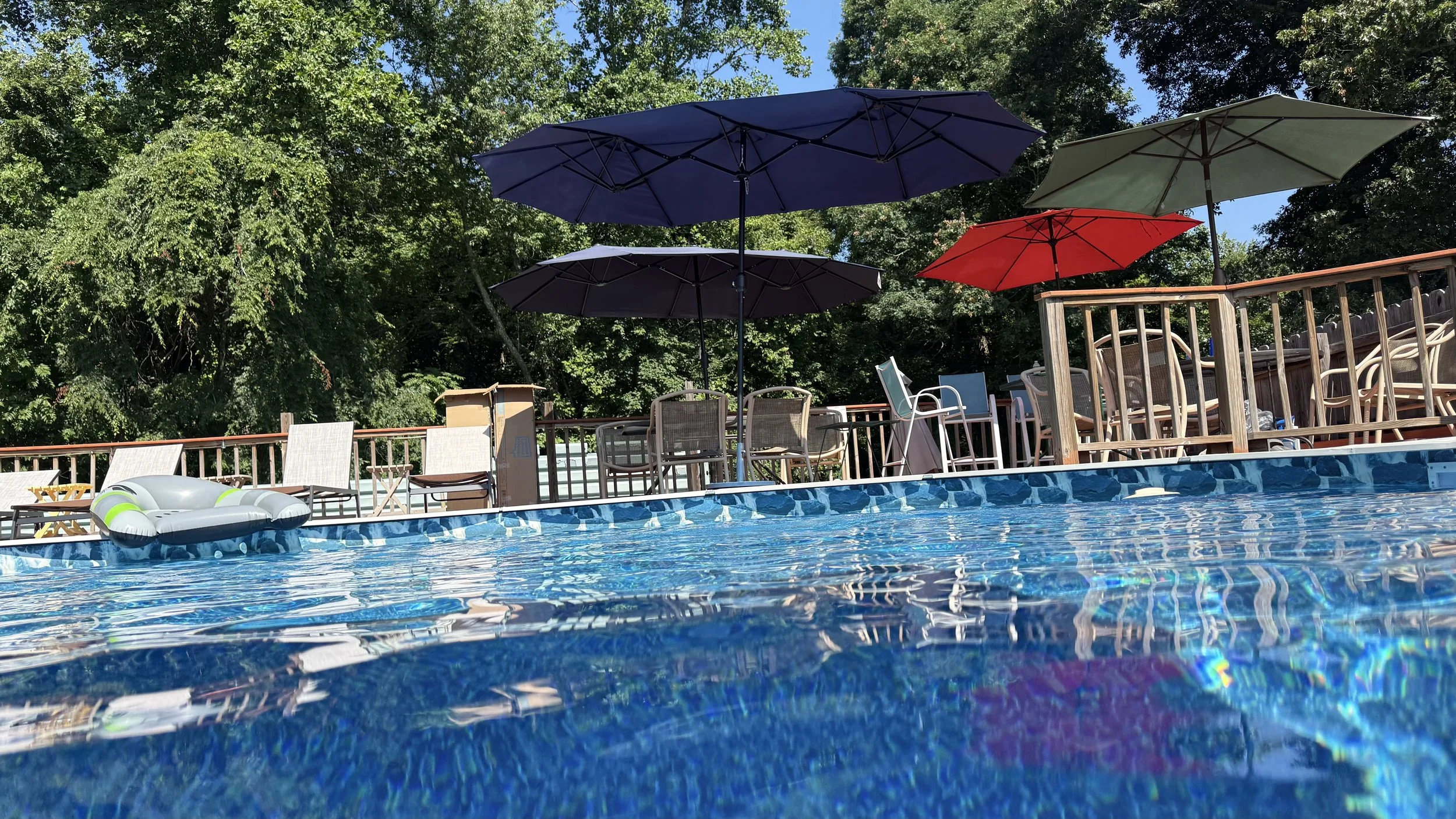 View of a swimming pool with blue water, surrounded by wooden deck chairs, umbrellas in various colors, and a lush green tree background on a sunny day.