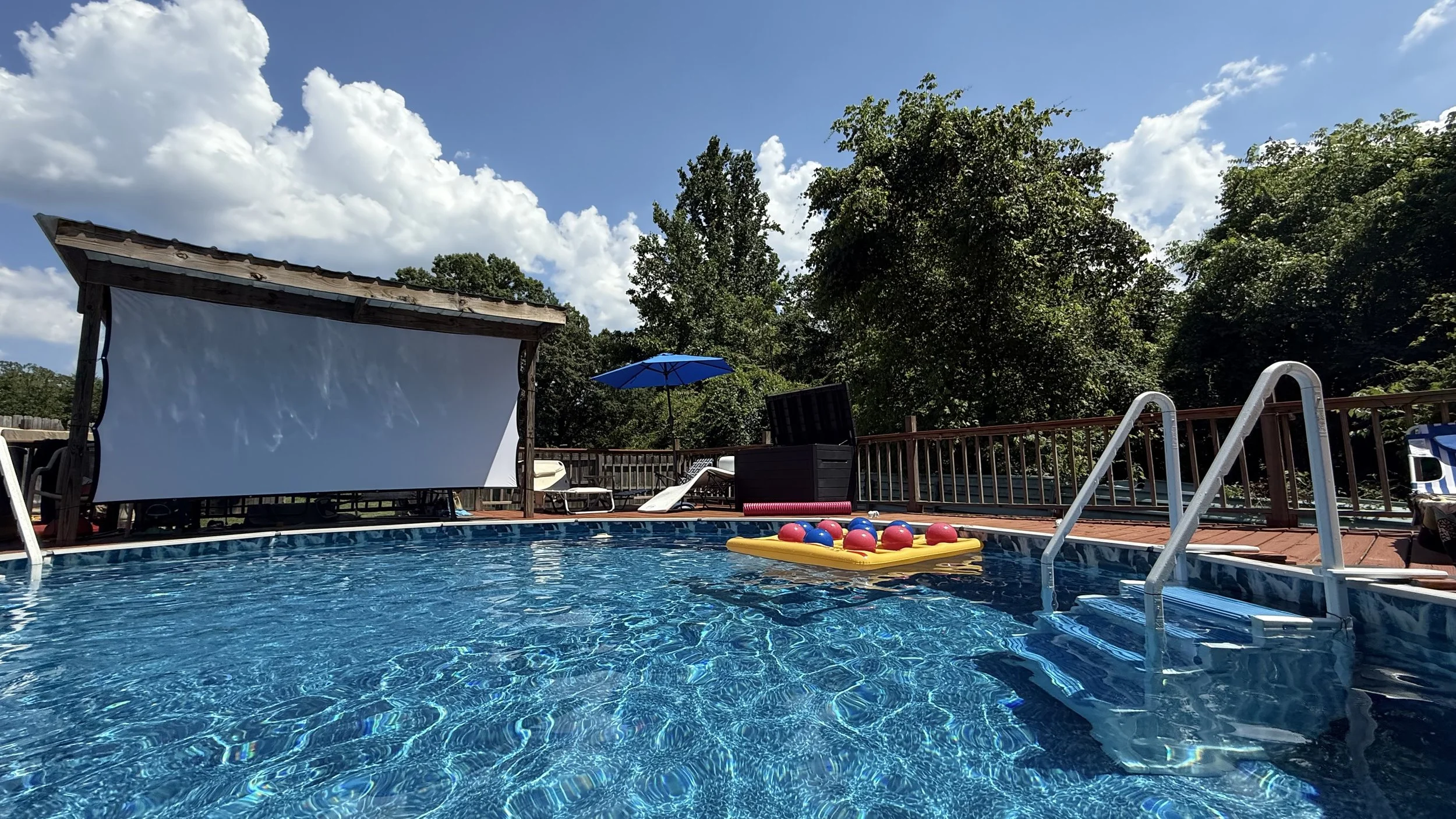 Swimming pool with an outdoor movie screen, pool toys, an umbrella, deck chairs, and lush green trees in the background under a partly cloudy sky.