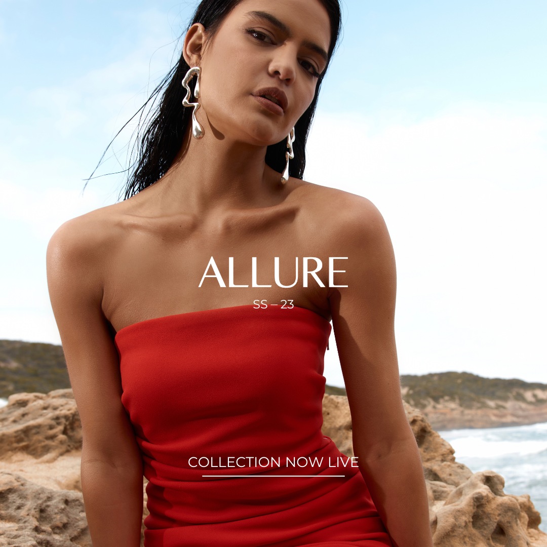 Model wearing a red strapless dress and silver earrings, standing on a rocky coastline with the text 'ALLURE SS-23 Collection Now Live'.