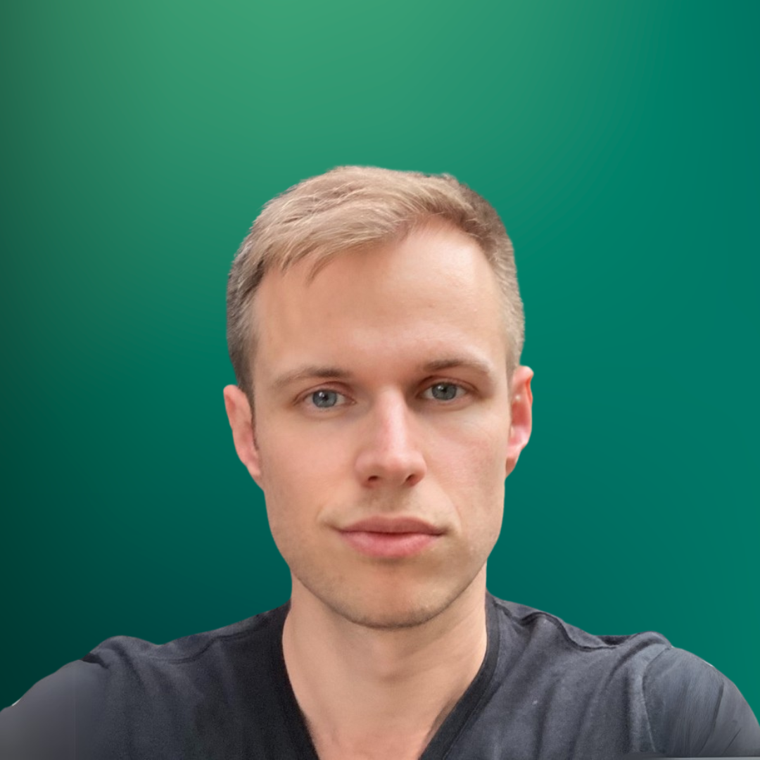 A young man with short blonde hair and blue eyes wearing a black shirt with a dark green background.