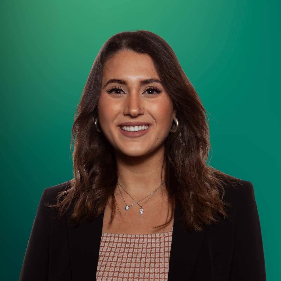 A smiling woman with shoulder-length brown hair, wearing a black blazer over a patterned top, with layered necklaces and hoop earrings, standing against a green gradient background.