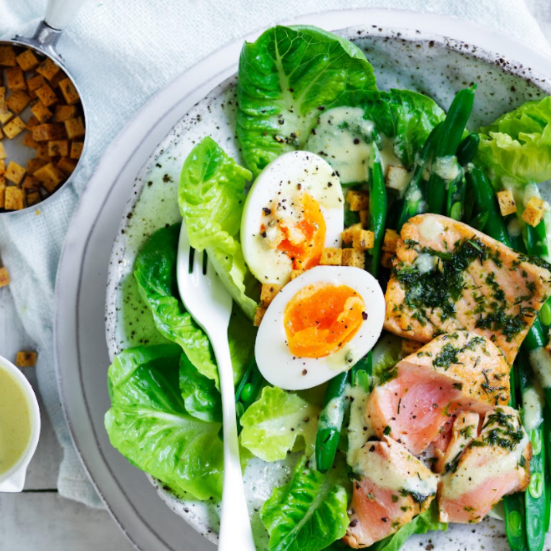 A salad with lettuce, boiled eggs, salmon, green beans, croutons, and dressing on a plate.