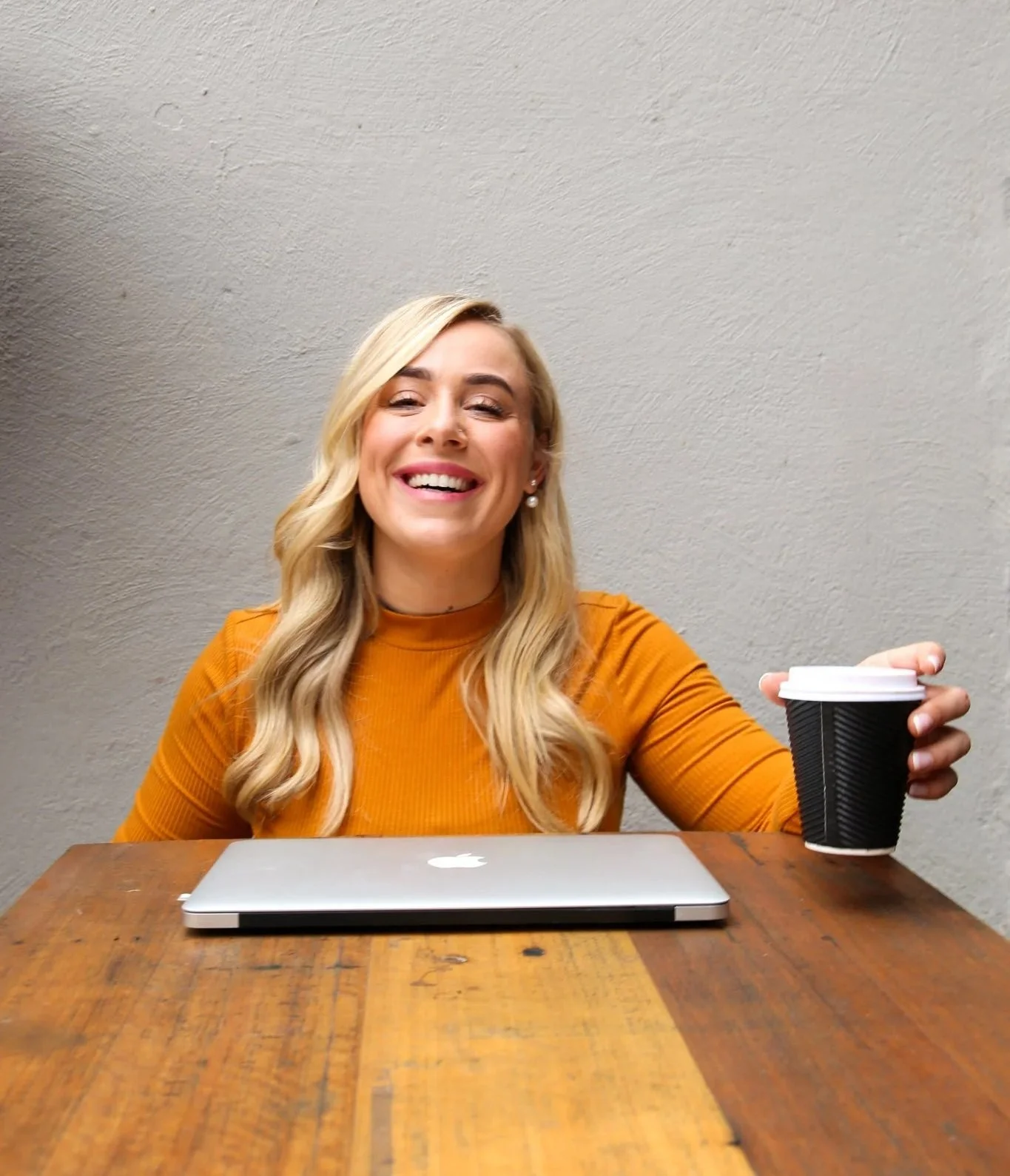 A woman with blonde hair, wearing an orange top, is sitting at a wooden table with a closed silver laptop and holding a black coffee cup, smiling at a camera against a plain gray wall.