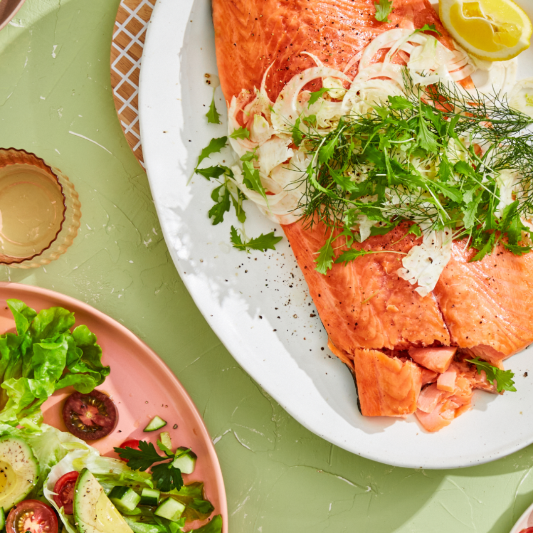 Cooked salmon fillet topped with herbs and lemon on a plate, next to a green salad with tomatoes, cucumbers, and avocado.