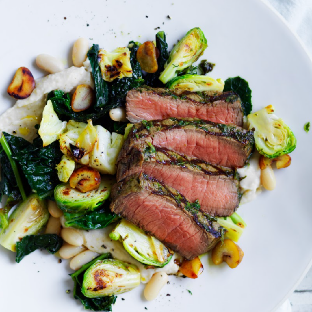 Sliced steak with Brussels sprouts, kale, roasted garlic, and white beans on a white plate.