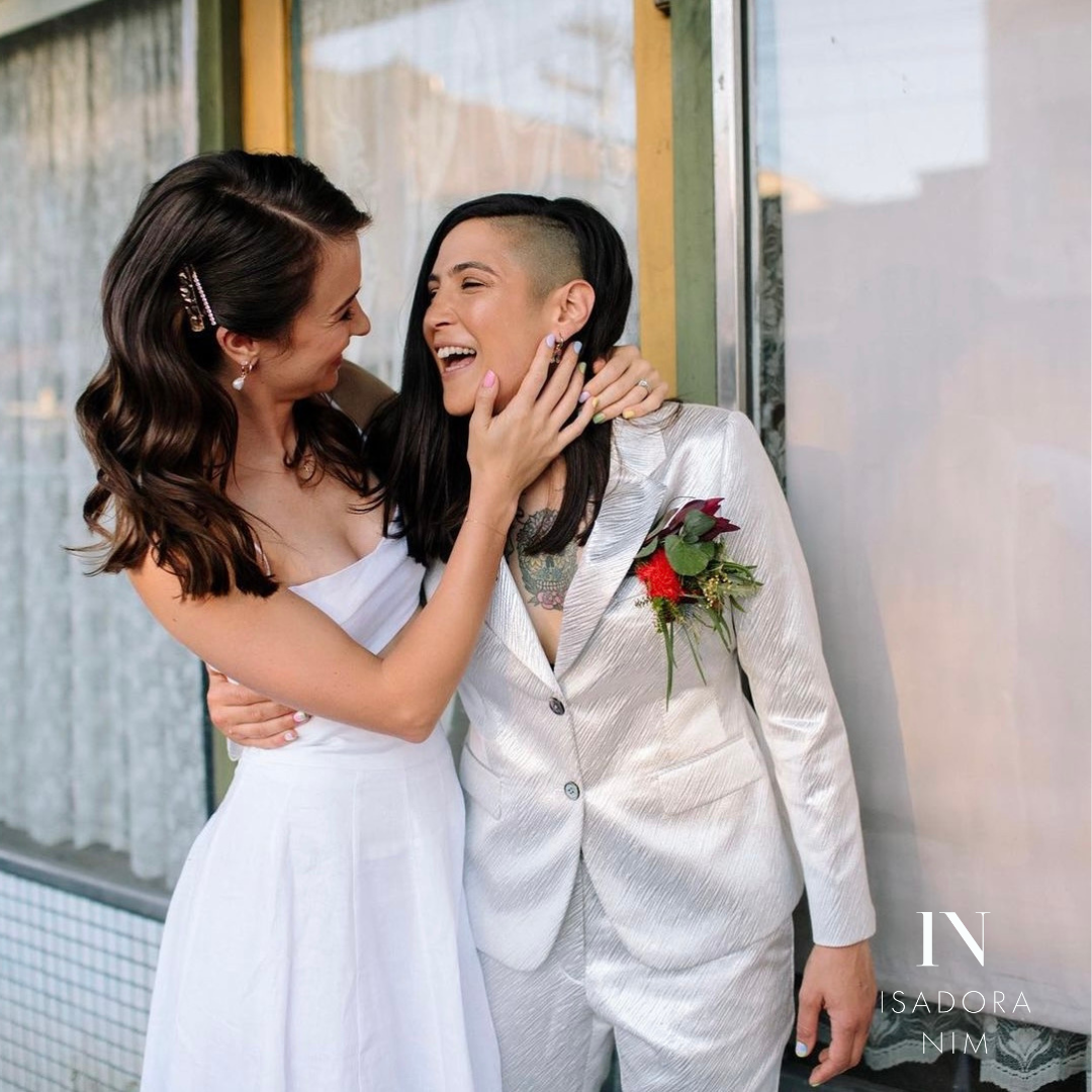 Two people joyfully embracing; one wearing a white dress, the other a light-colored suit with a boutonniere.