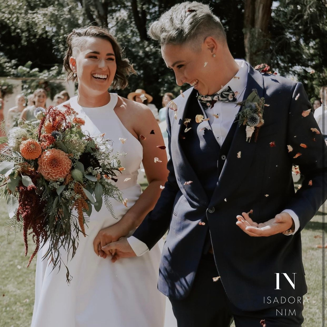 Two people smiling and holding hands, dressed in wedding attire with flower petals falling around them. One is wearing a white gown and holding a bouquet, while the other is in a dark suit. They are outdoors with greenery in the background.
