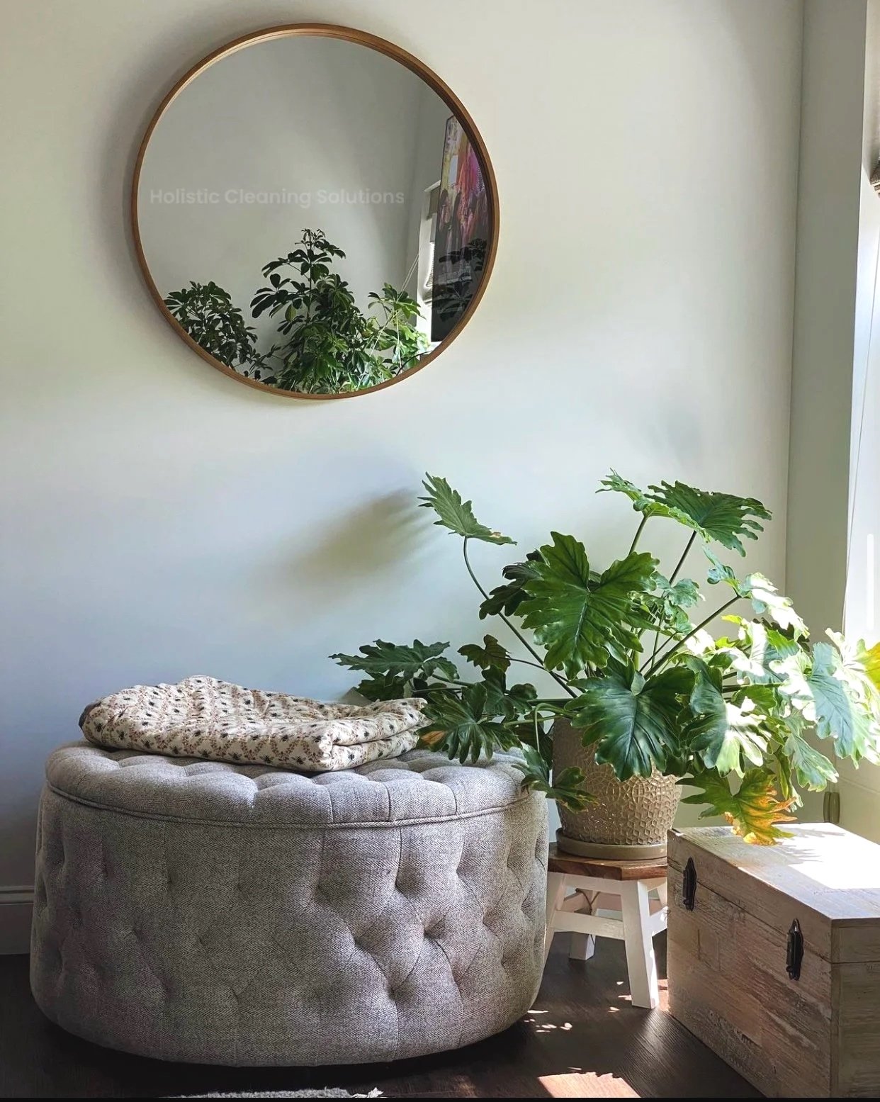 Living room corner with a large round upholstered ottoman, a potted plant with big green leaves, a vintage wooden chest, a small white stool, a wall mirror, and sunlight streaming in.