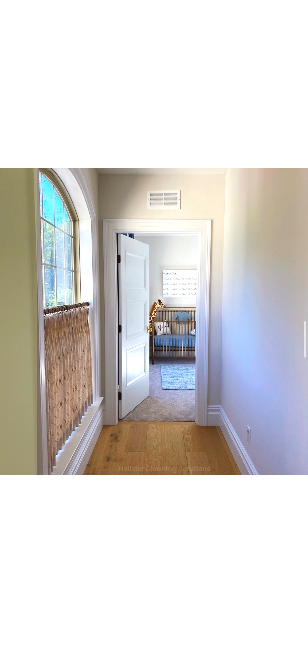 A hallway with hardwood flooring and light-colored walls leading to a nursery room with a crib, a giraffe plush toy, and a hand-written wall decor. Sunlight streams through an arched window on the left with a wooden window treatment.