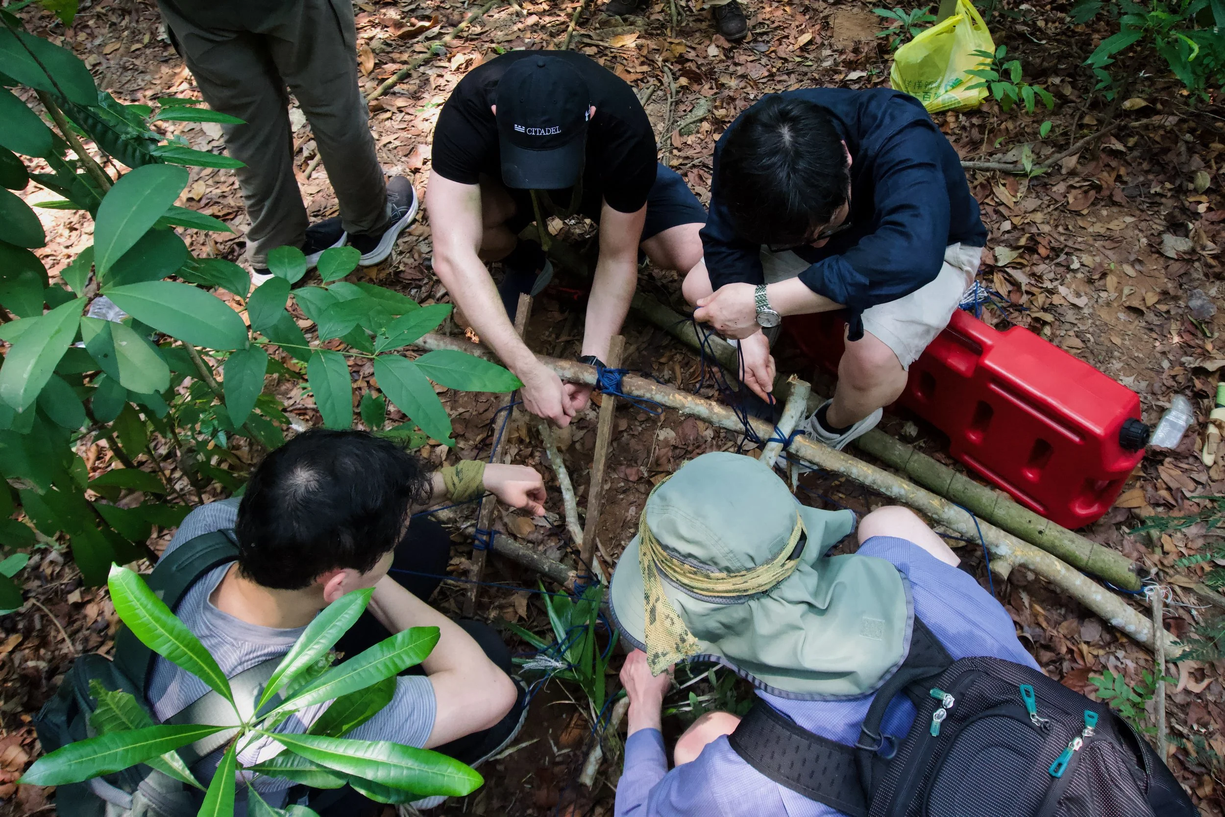 Citadel Securities participants lashing wooden poles together with rope to build an improvised stretcher, with a red fuel can beside them.