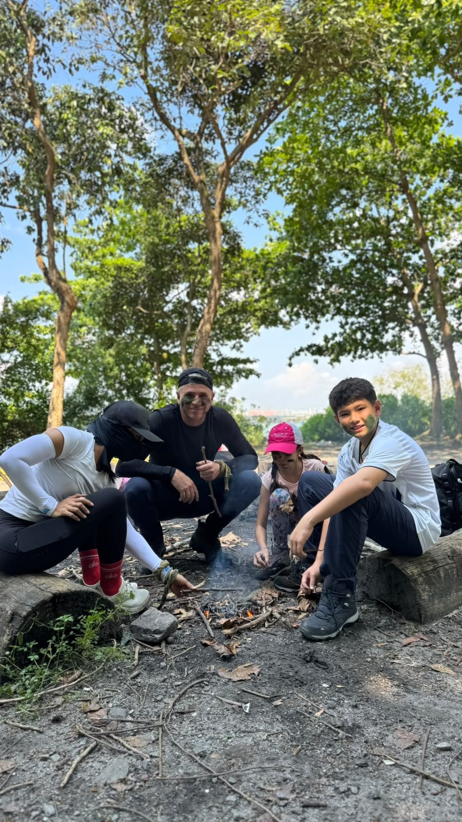 Family sitting on logs around a small fire with their guide in a Pulau Ubin clearing