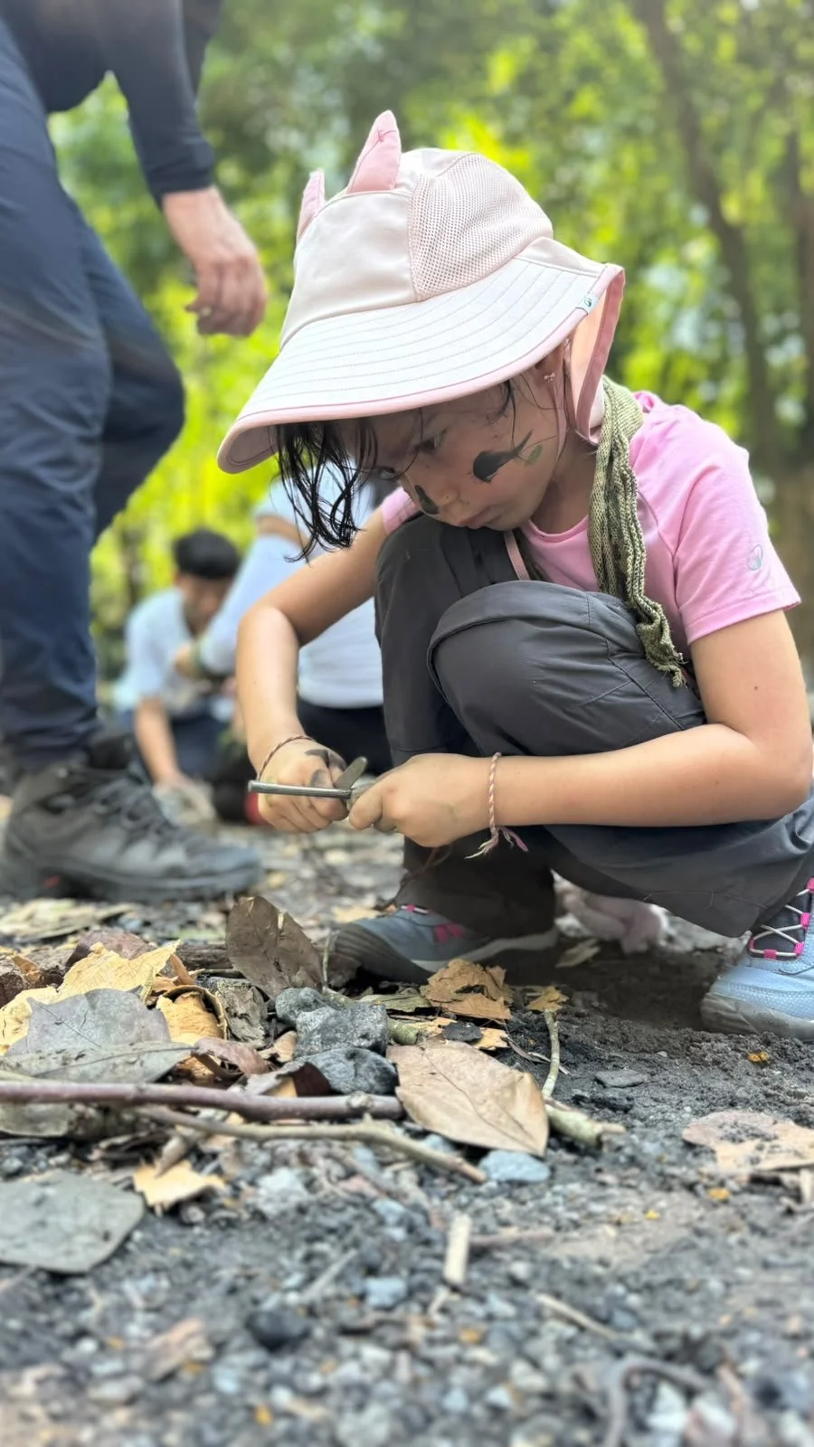 Girl in a white sun hat crouching low to strike a ferro rod over dry leaves and tinder
