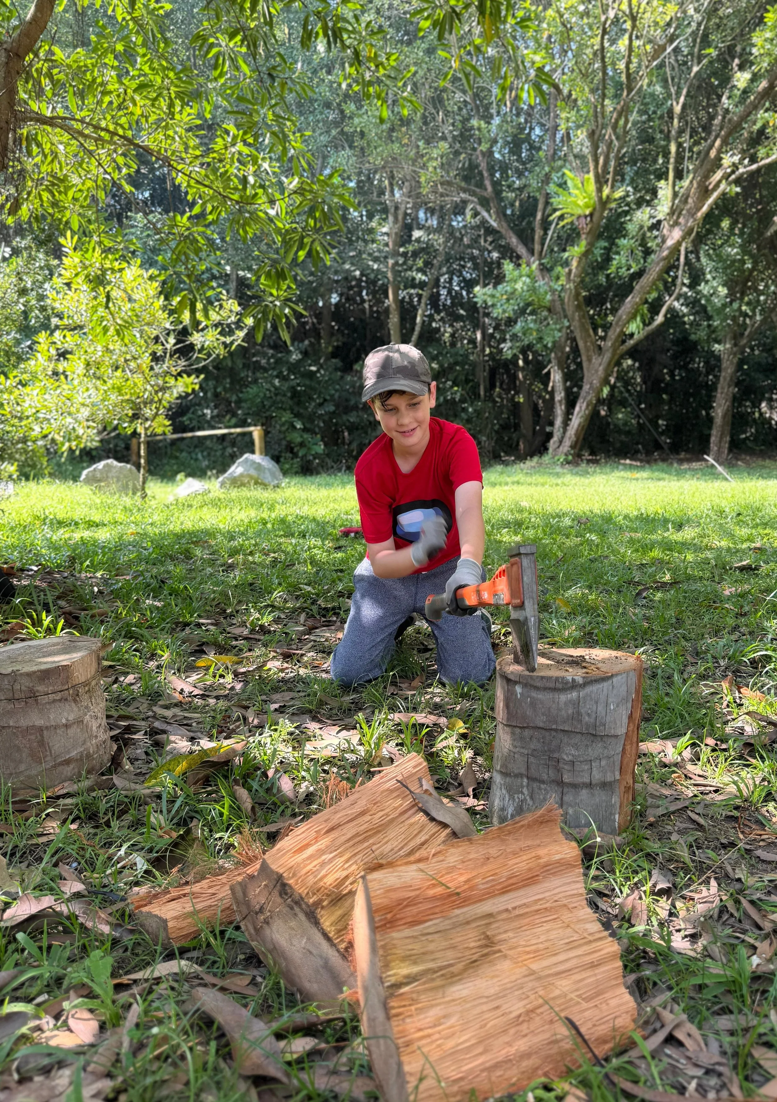Boy in a red shirt kneeling on grass with a hatchet beside a chopping block and split firewood