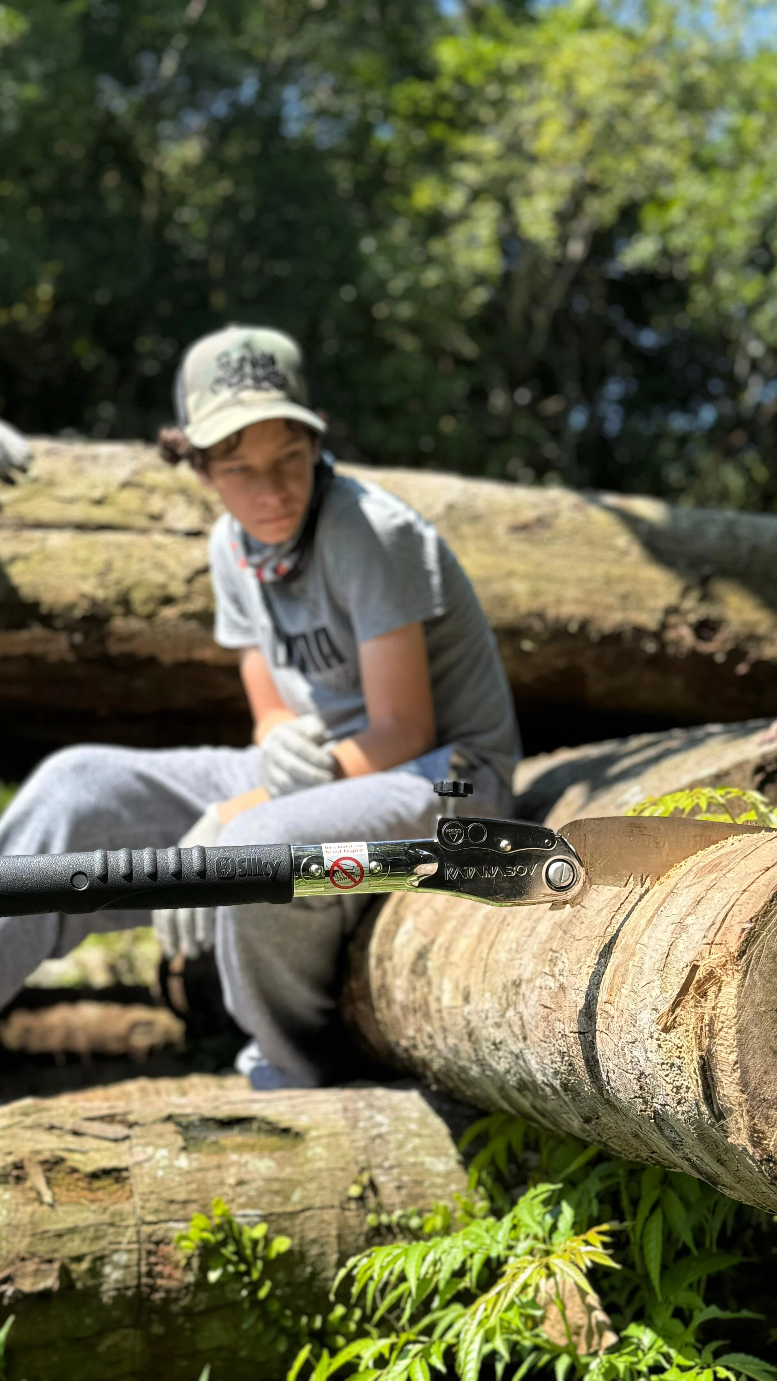 Teenager in gloves and a cap sitting on a felled log while cutting it with a large saw