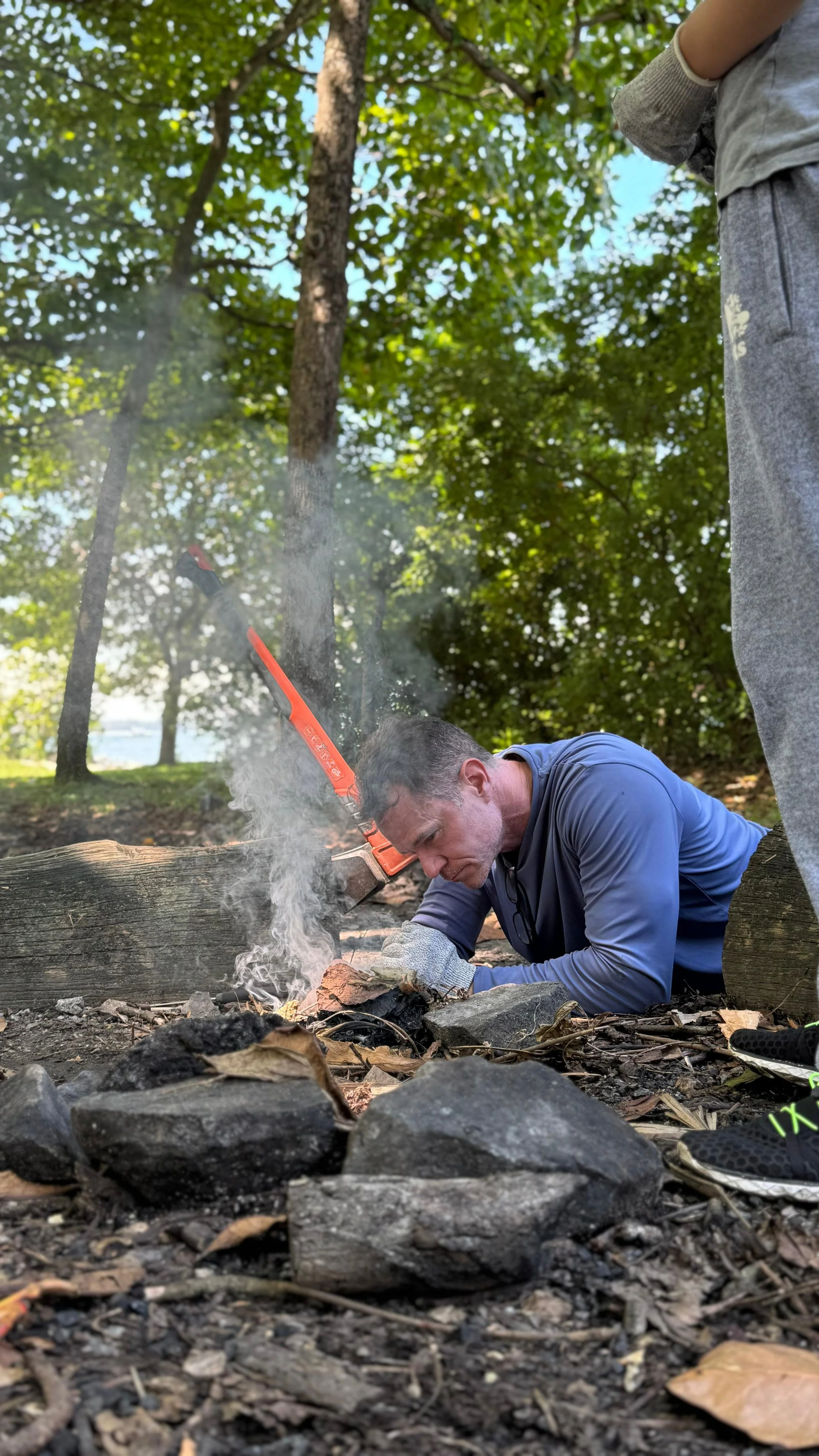 Man kneeling low to blow into a smoking fire ringed with stones as another person stands nearby