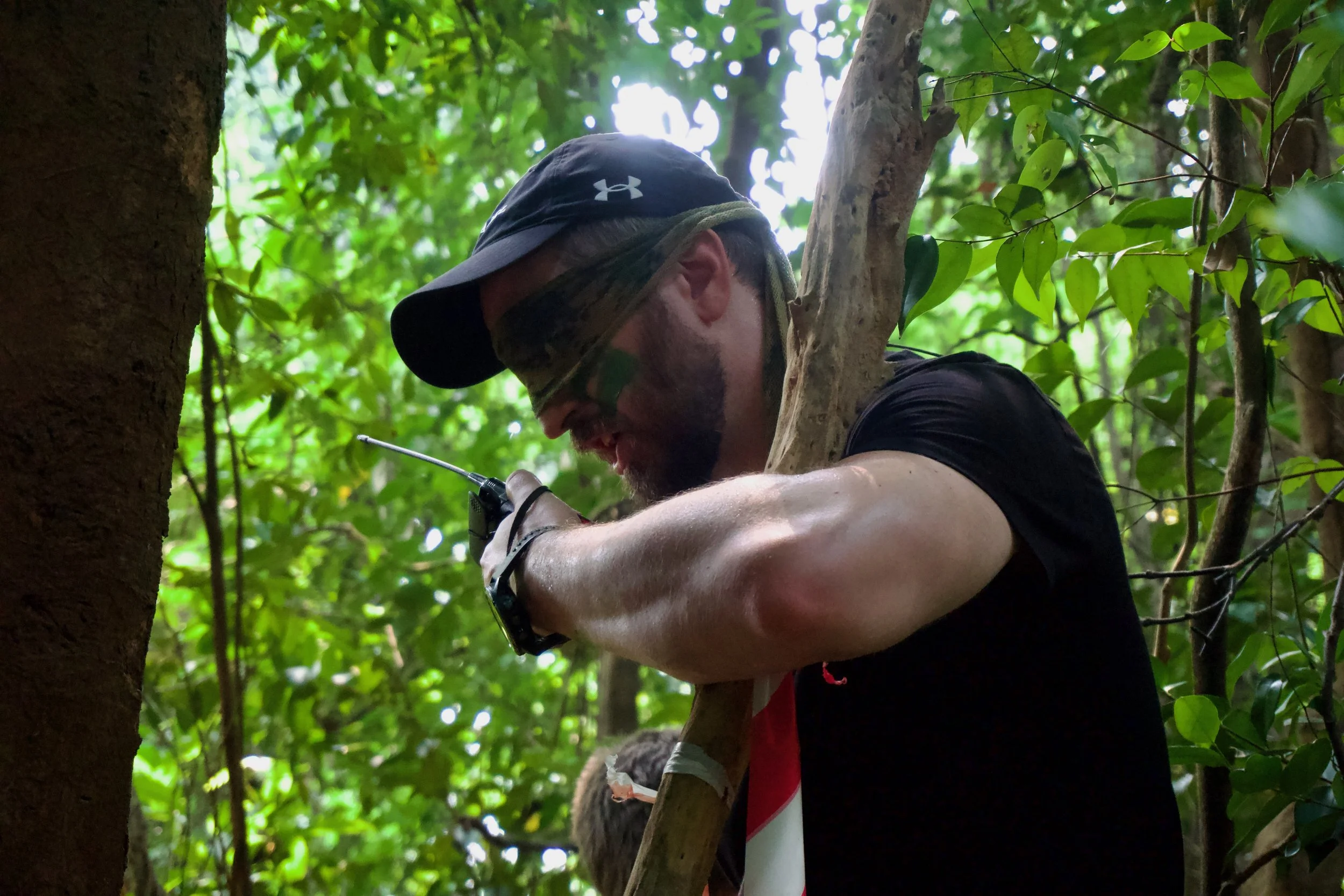 Participant with camouflage face paint steadying himself against a tree in dense forest