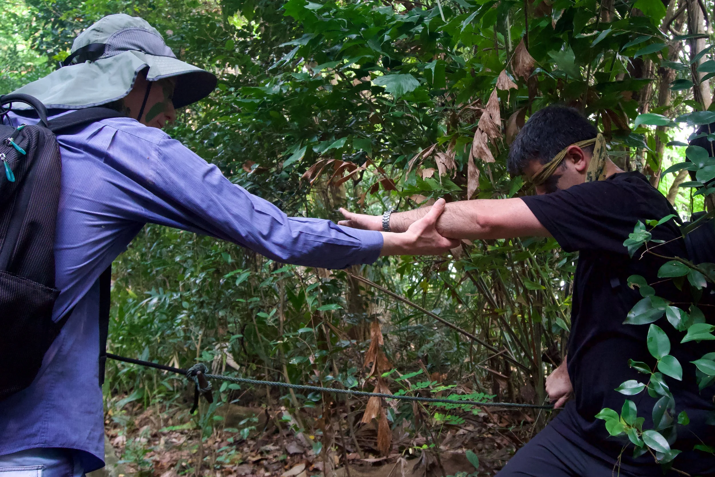 Two participants reaching for each other along a guide rope in forest terrain