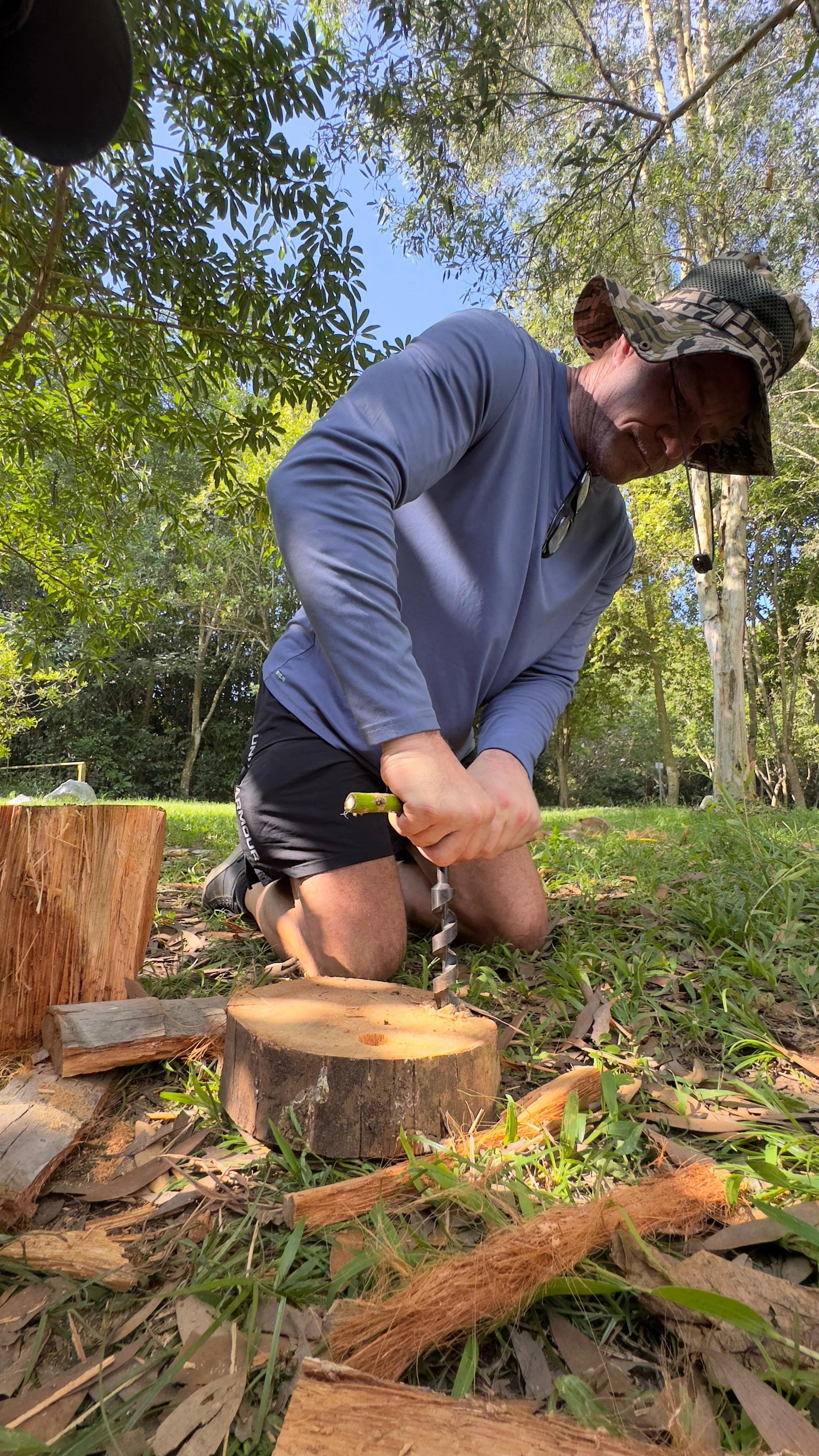 Father kneeling on grass turning a manual auger to bore a hole through a log round.
