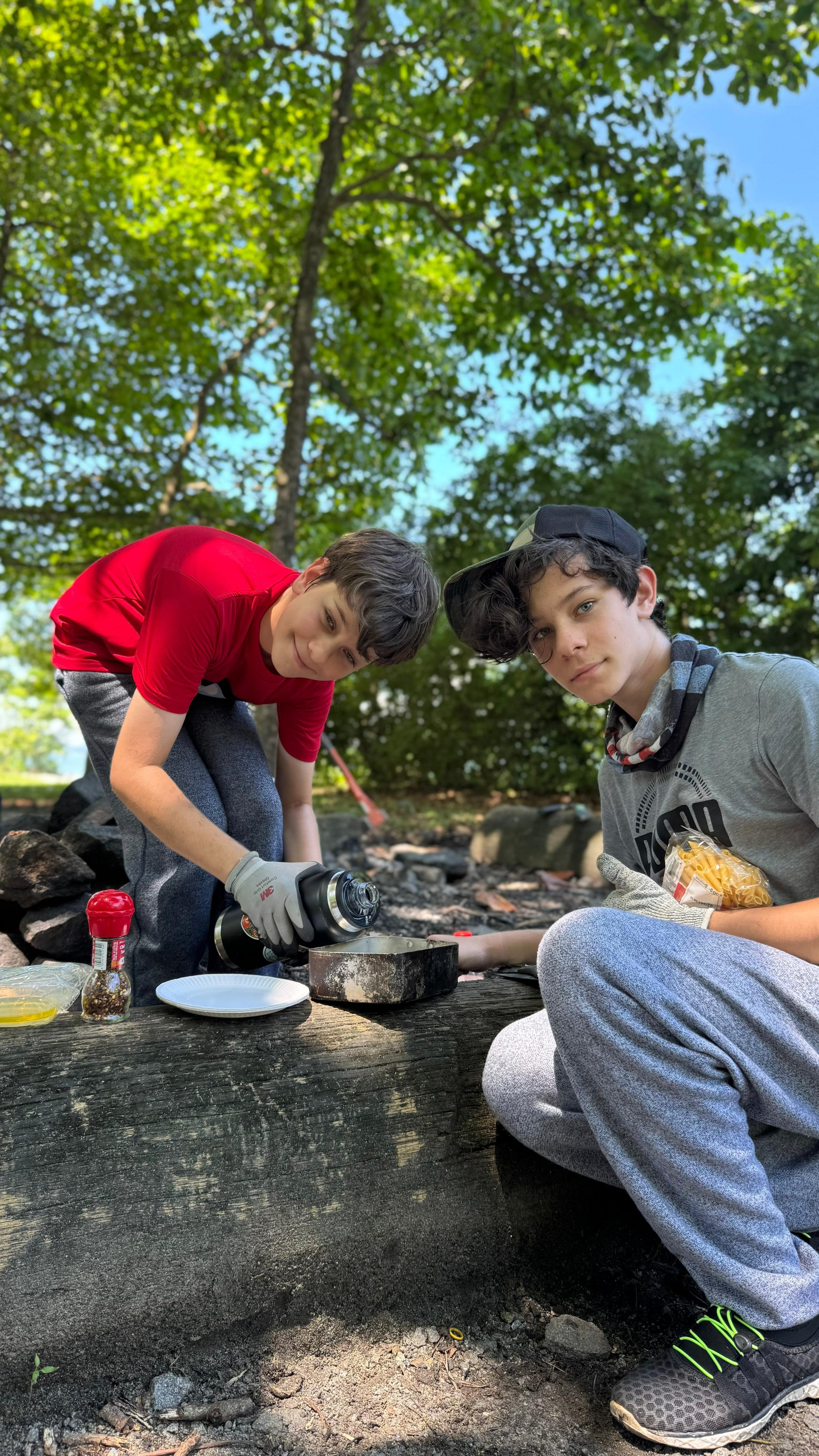 Two boys crouching beside a fire pit, pouring water into a metal mess tin with noodles and seasoning beside them.