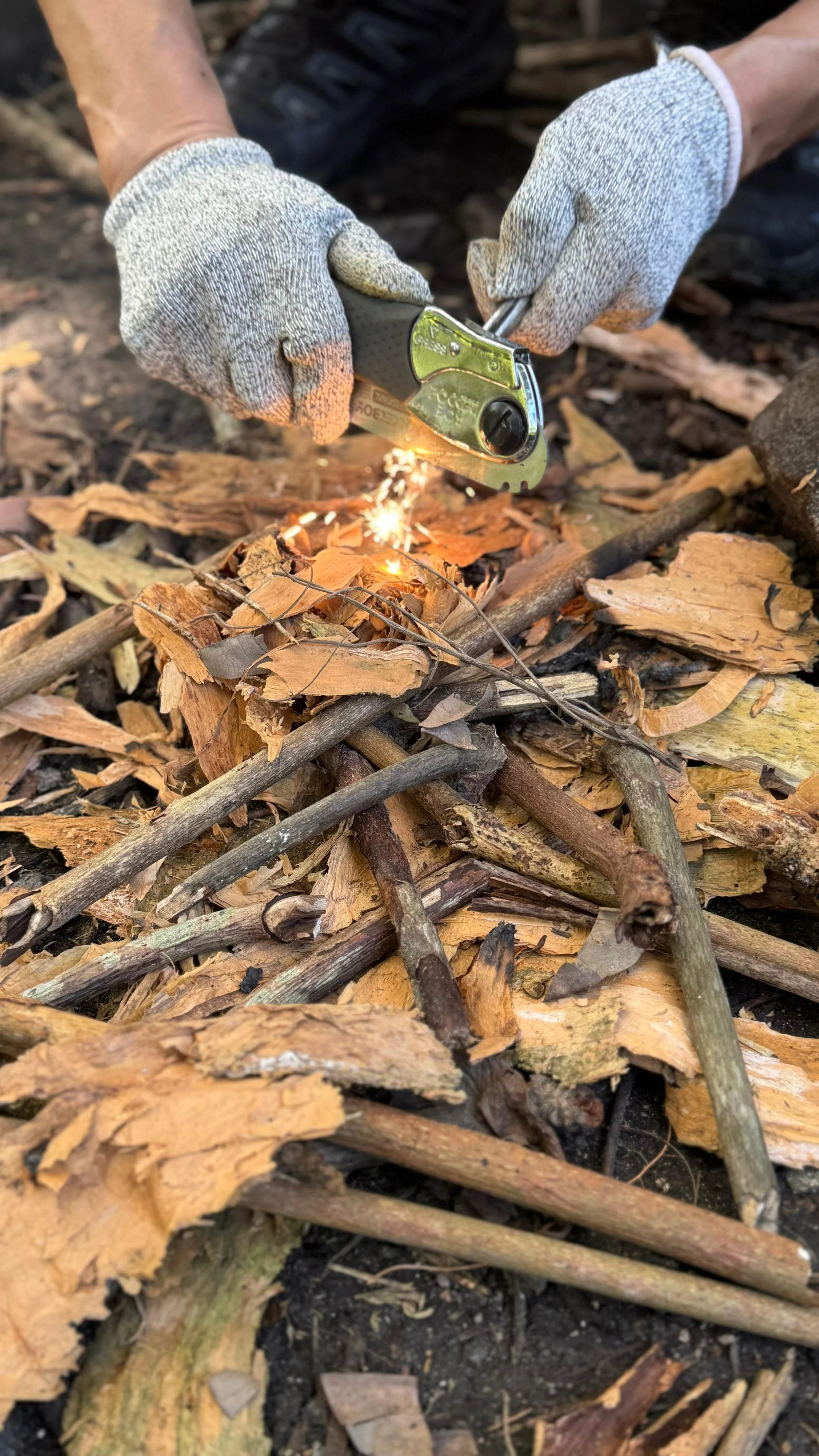 Gloved hands striking a ferro rod, sending sparks onto a prepared bed of dry bark and twigs.