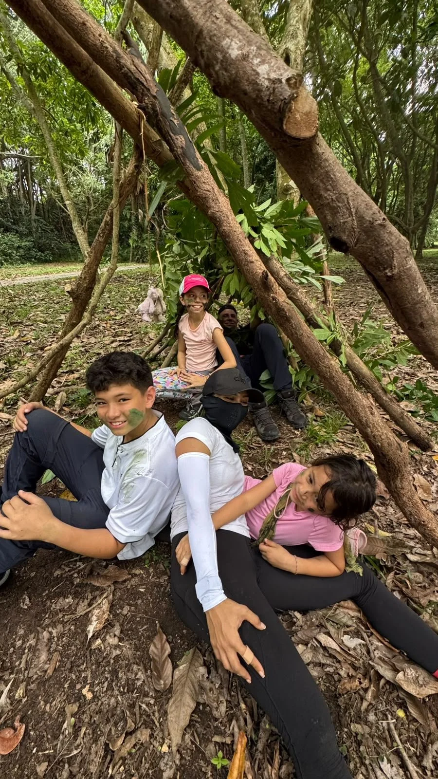 Family sitting under the A frame branch shelter they built together in the woods on Pulau Ubin