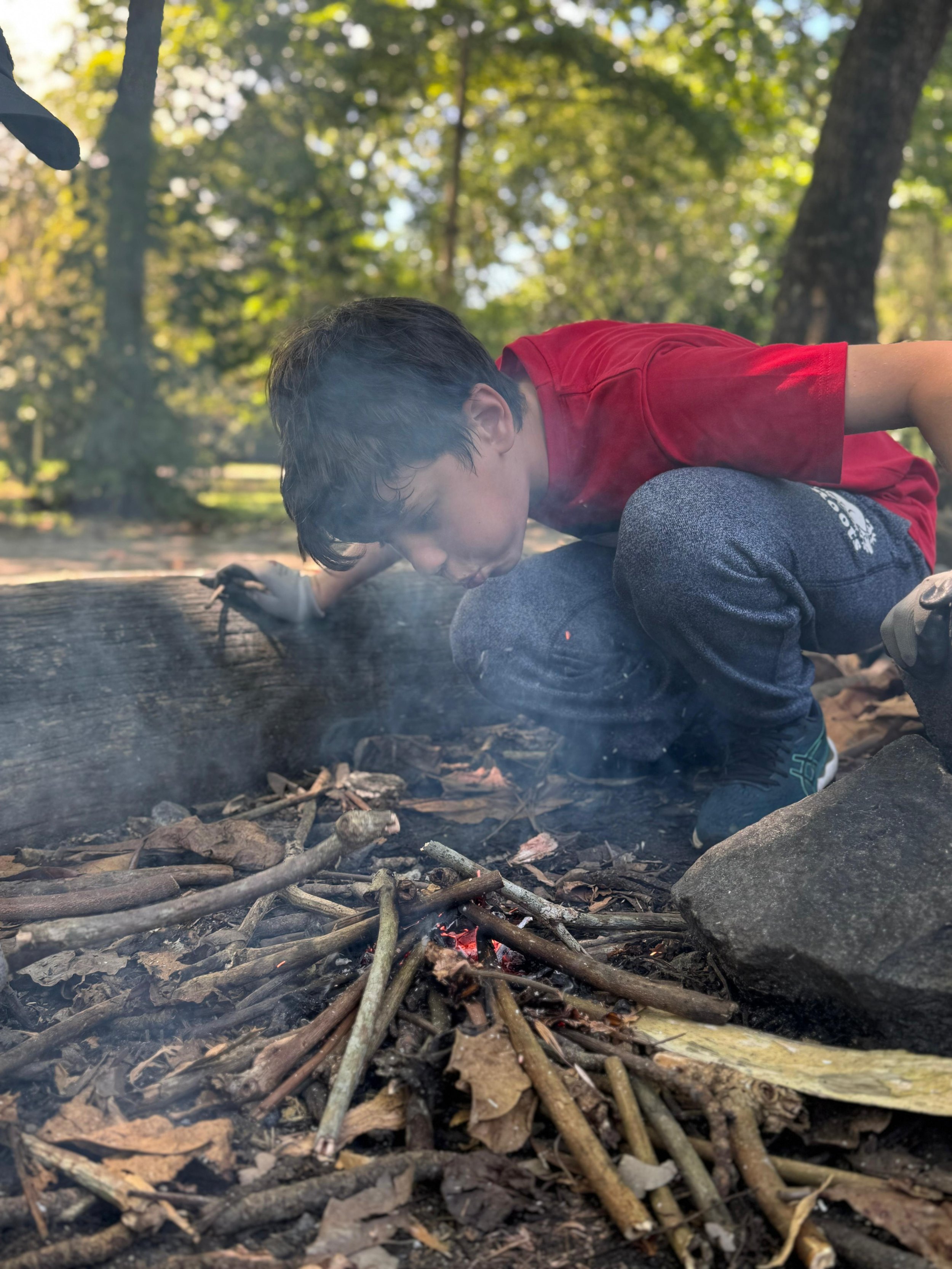 Boy crouching low to the ground, blowing steadily on a smoking bundle of sticks and leaves.
