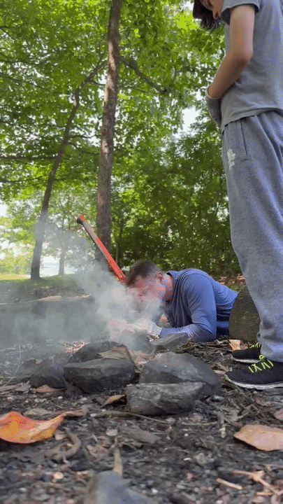 Father lying on the ground blowing into a smoking fire as thick smoke rises around him.