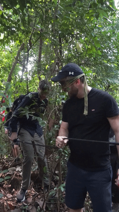 Blindfolded Citadel Securities participants stepping over a low obstacle in dense forest terrain during the blind infiltration at Fort Serapong.
