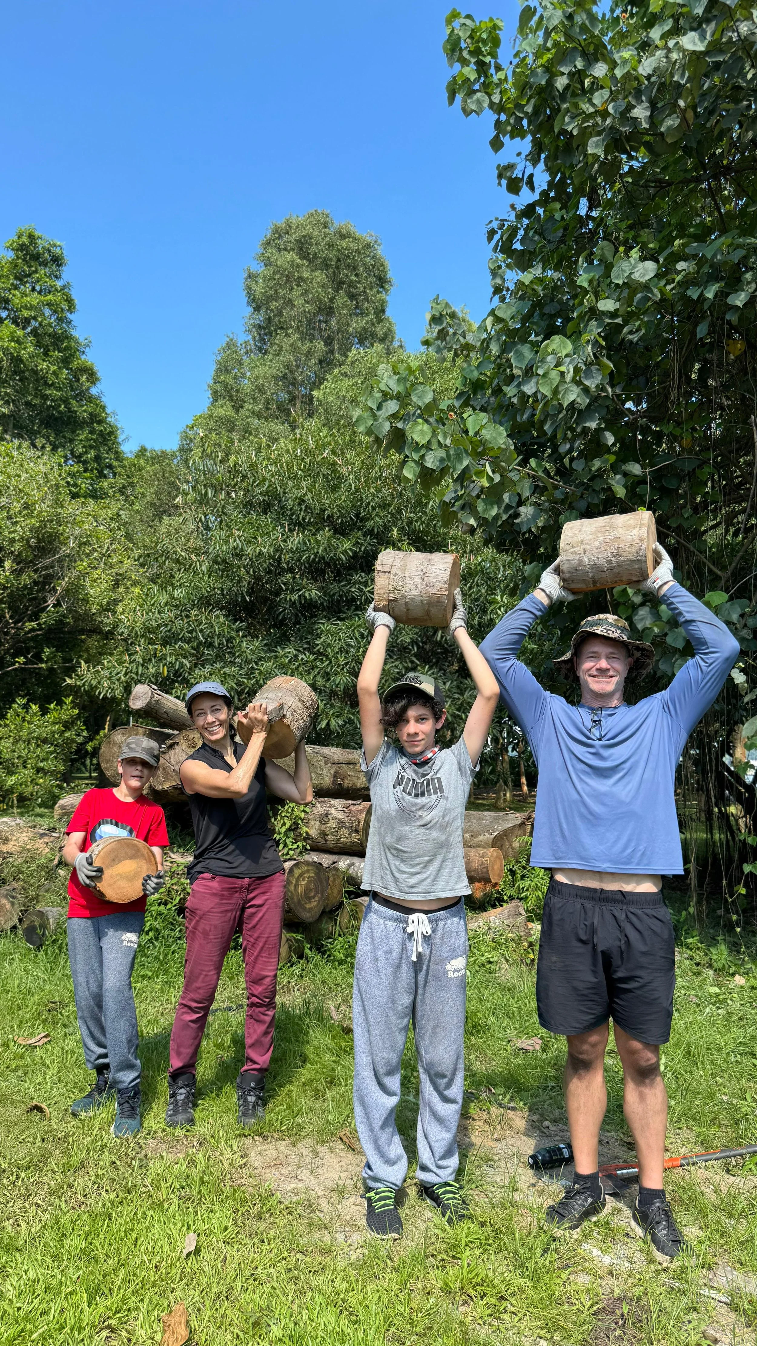 Family of four standing beside a log pile, holding freshly cut timber rounds after a bushcraft day on Pulau Ubin.