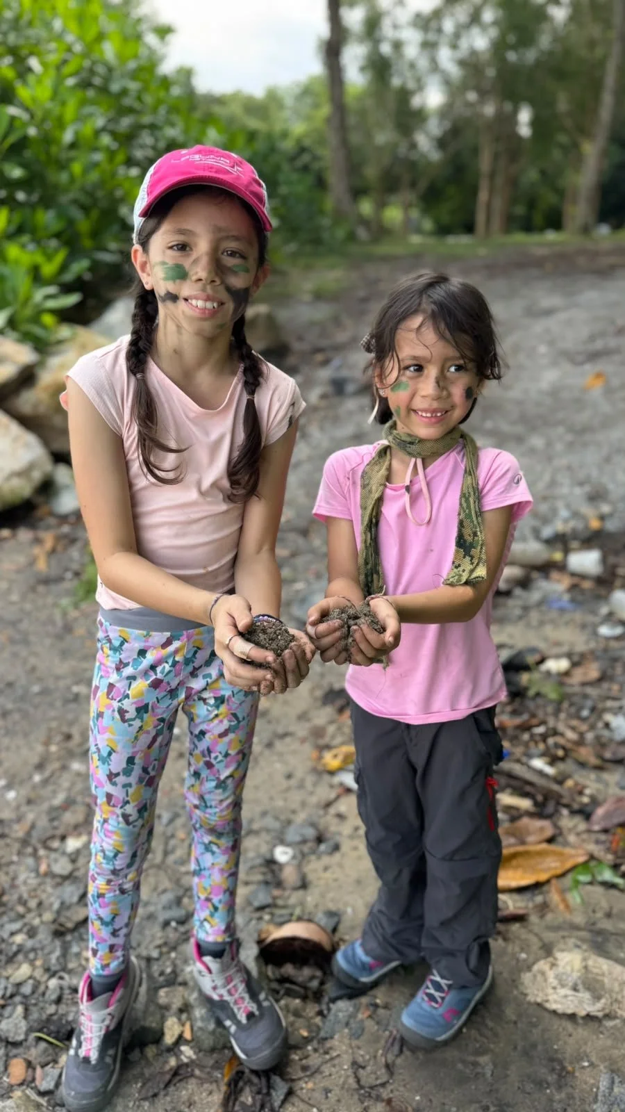 Two young sisters with face paint holding up handfuls of wet mud on a gravel track on Pulau Ubin