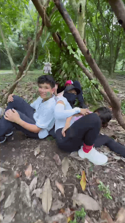 Three siblings sitting and lying inside the finished branch shelter on the forest floor at Pulau Ubin
