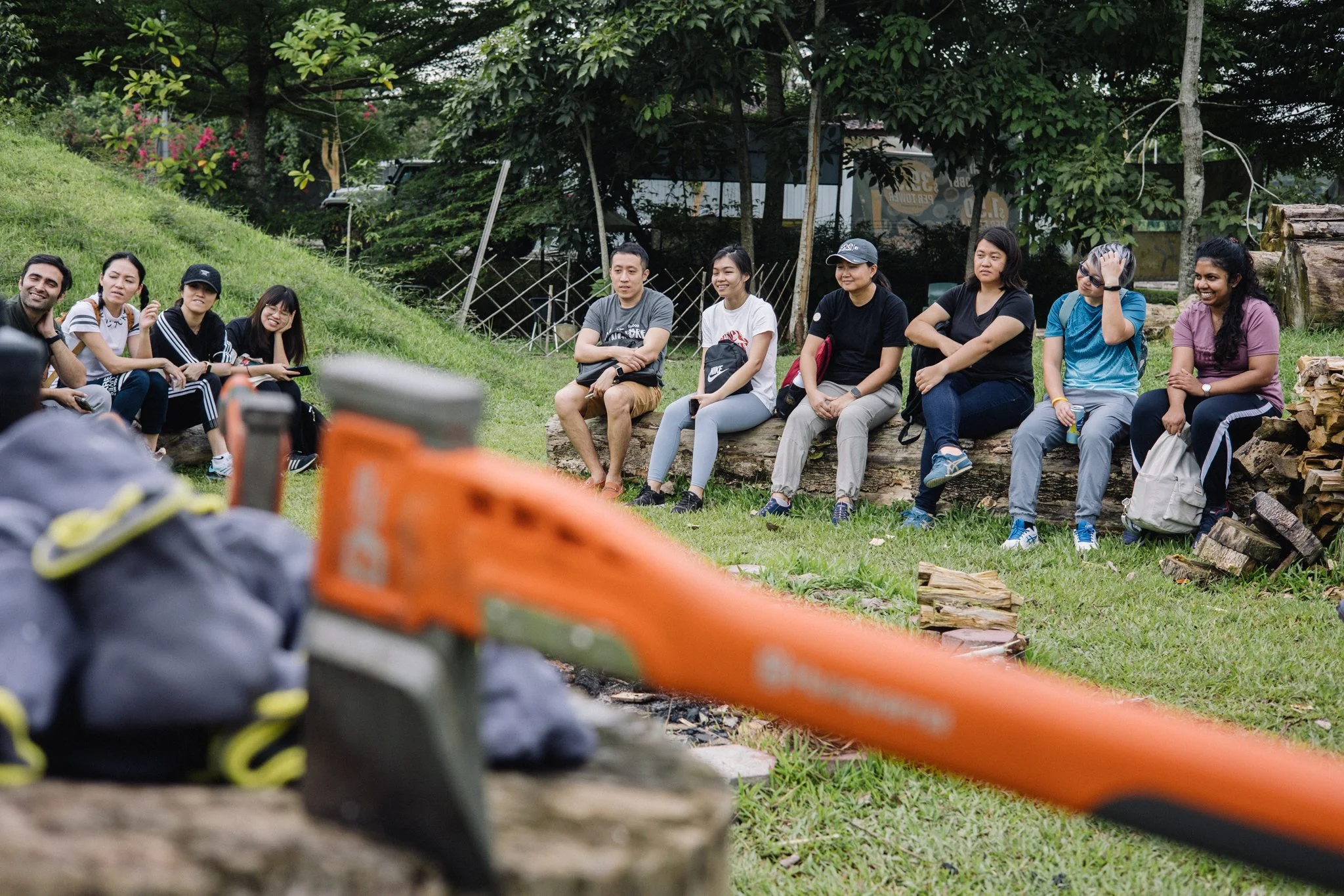 A Singapore corporate group seated on logs during an outdoor team bonding briefing, with a splitting axe in the foreground.