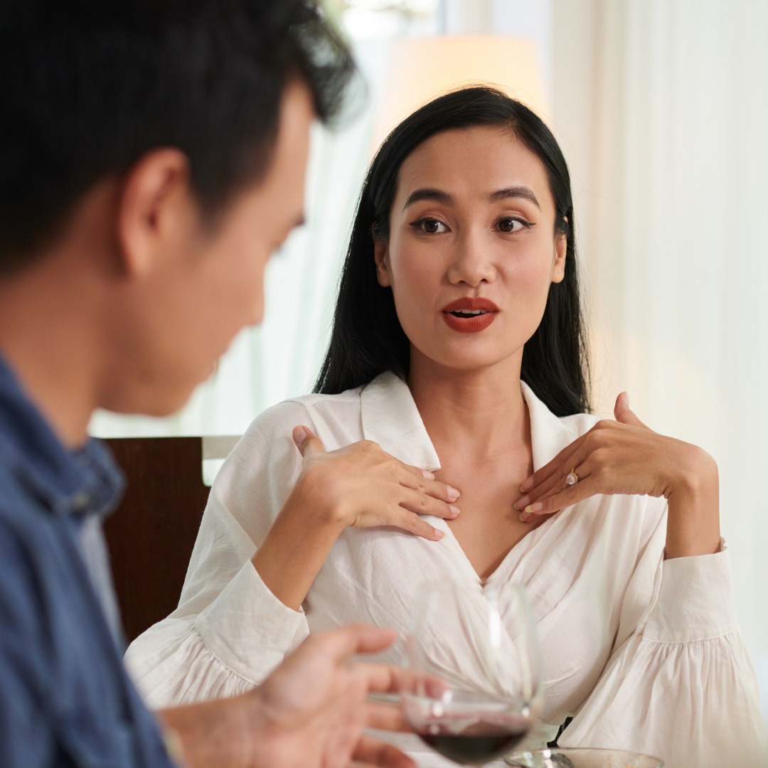 A woman with long black hair and a white blouse speaking, with her hands on her chest, while a man with short hair, wearing a navy shirt, is holding a glass of red wine and listening.