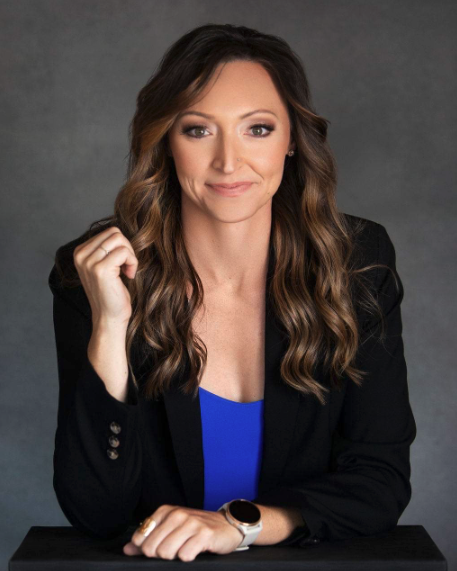 A woman with long wavy brown hair wearing a black blazer and blue top, sitting against a gray background.