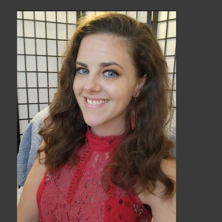A woman with long, curly brown hair and blue eyes smiling, wearing a red lace top and red earrings, in front of a grid-patterned background.