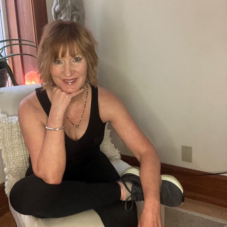 A woman with shoulder-length reddish hair sitting on a white cushioned chair, smiling, with her chin resting on her hand. She is wearing a black sleeveless top, black pants, and sneakers, in a room with a beige wall and wooden floor.