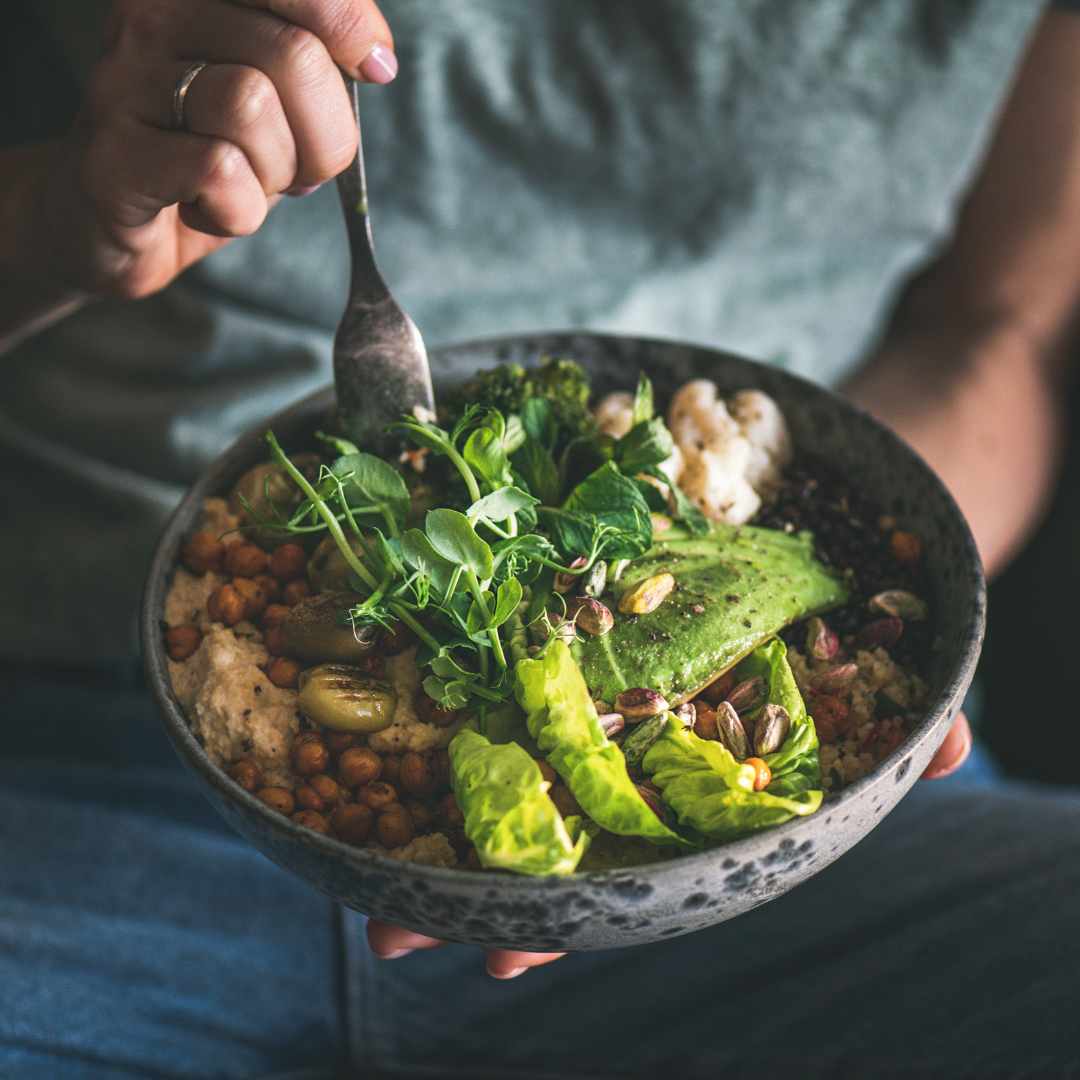A person holding a black bowl filled with fresh salad greens, avocado slices, cauliflower, chickpeas, and other vegetables, using a fork.