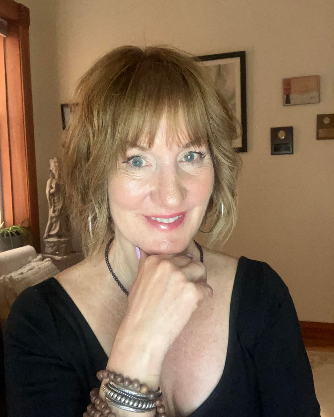 A woman with shoulder-length reddish-brown hair and blue eyes, smiling and resting her chin on her hand, wearing a black top, jewelry including a necklace, bracelets, and earrings, in a cozy home setting with framed artwork on the wall behind her.