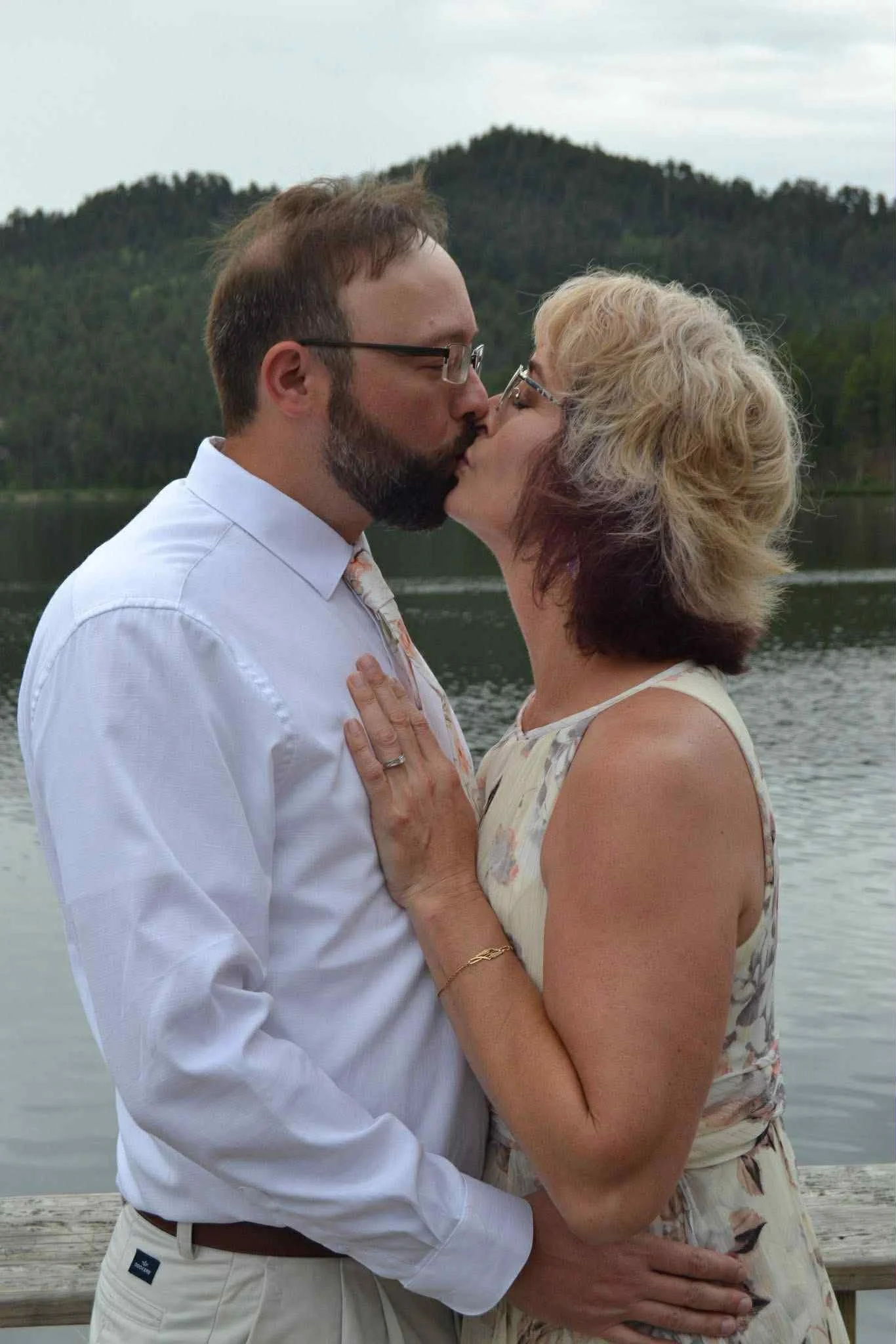 A couple kissing outdoors by a lake with mountains in the background.