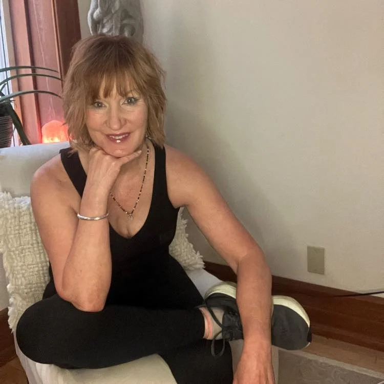 A woman with shoulder-length reddish hair sitting on a white cushioned chair, smiling, with her chin resting on her hand. She is wearing a black sleeveless top, black pants, and sneakers, in a room with a beige wall and wooden floor.
