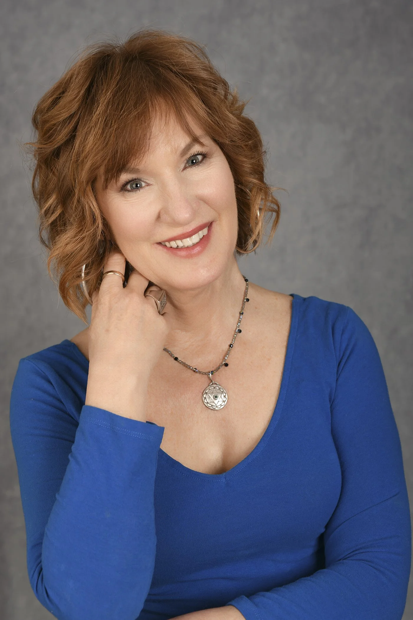 A woman with shoulder-length curly red hair smiling, wearing a blue top, a black and silver bead necklace, and a silver pendant necklace, against a gray background.
