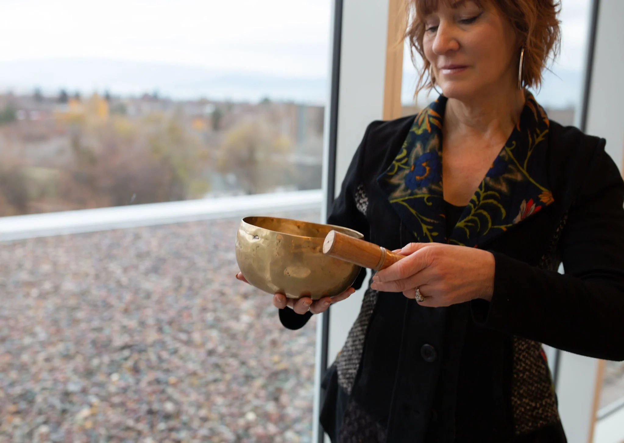 A woman holding a singing bowl and a wooden striker, standing indoors near a window with a view of a cloudy, autumn landscape.