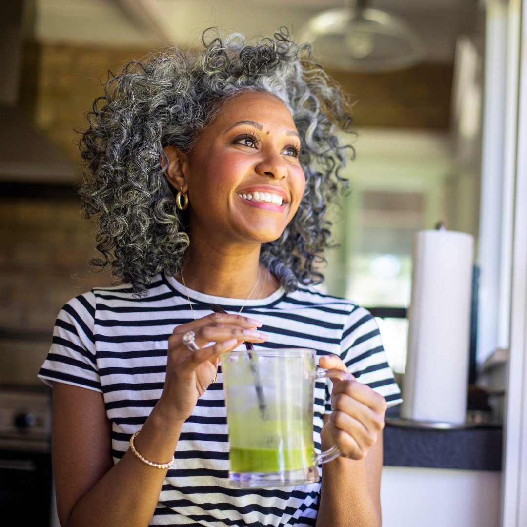 A smiling woman with gray curly hair wearing a black and white striped shirt, holding a glass of green smoothie with a straw, standing in a bright kitchen.