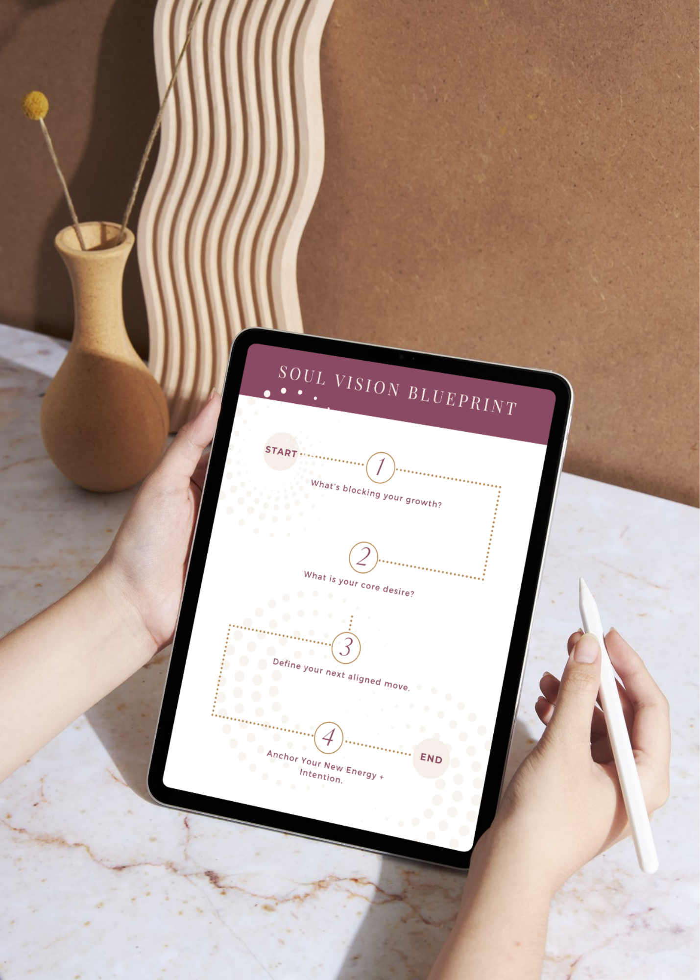 A person holding a tablet with a 'Soul Vision Blueprint' worksheet displayed on the screen. The worksheet has four steps, including prompts about growth, core desire, next move, and anchoring energy. The person also holds a white stylus. The background includes a decorative tan vase with two yellow spherical flowers and a wavy wooden sculpture against a corkboard wall, all on a white marble tabletop.