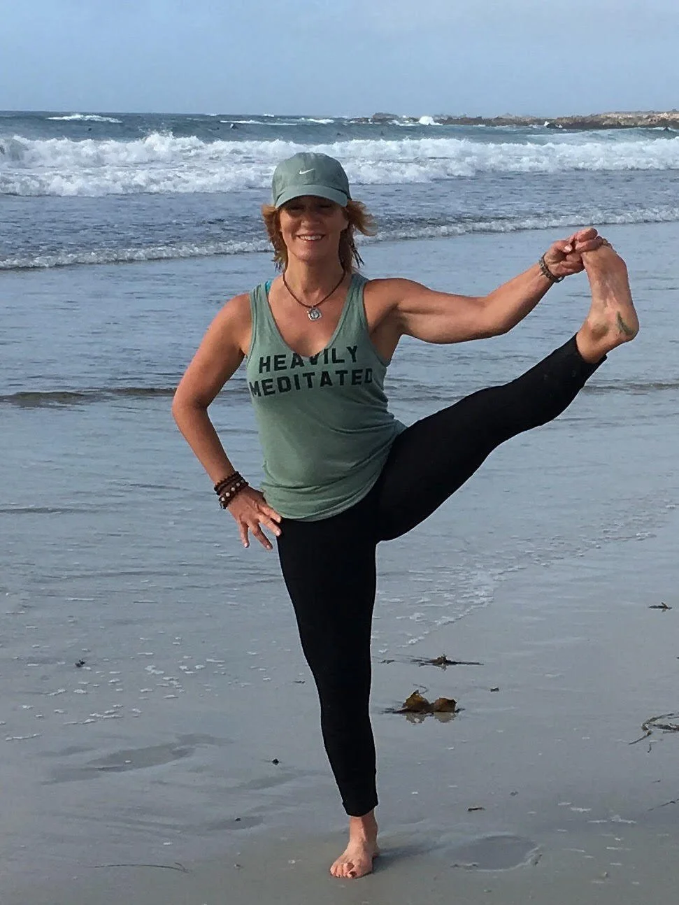 Laura Bender doing a yoga pose on the beach, wearing a green tank top with the words "HEAVILY MEDITATED," black leggings, a gray cap, and jewelry, with the ocean and waves in the background.