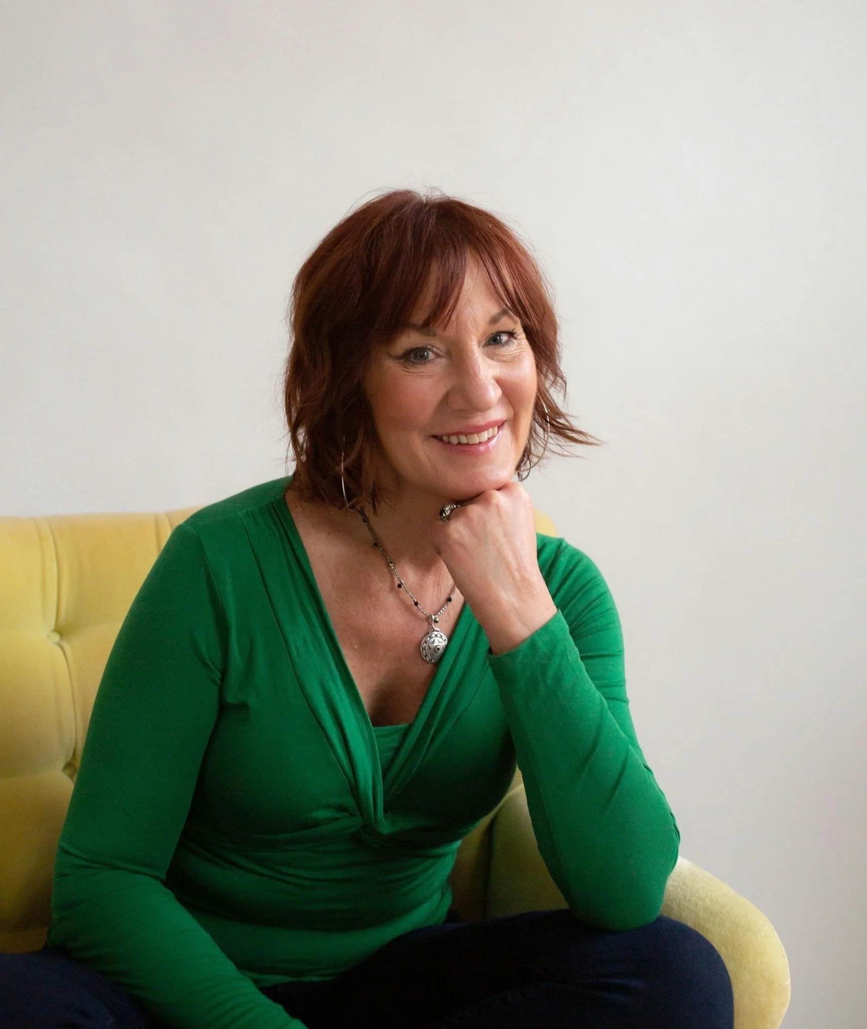 A woman with reddish-brown hair, wearing a green top, sitting on a yellow couch, smiling and resting her chin on her hand.