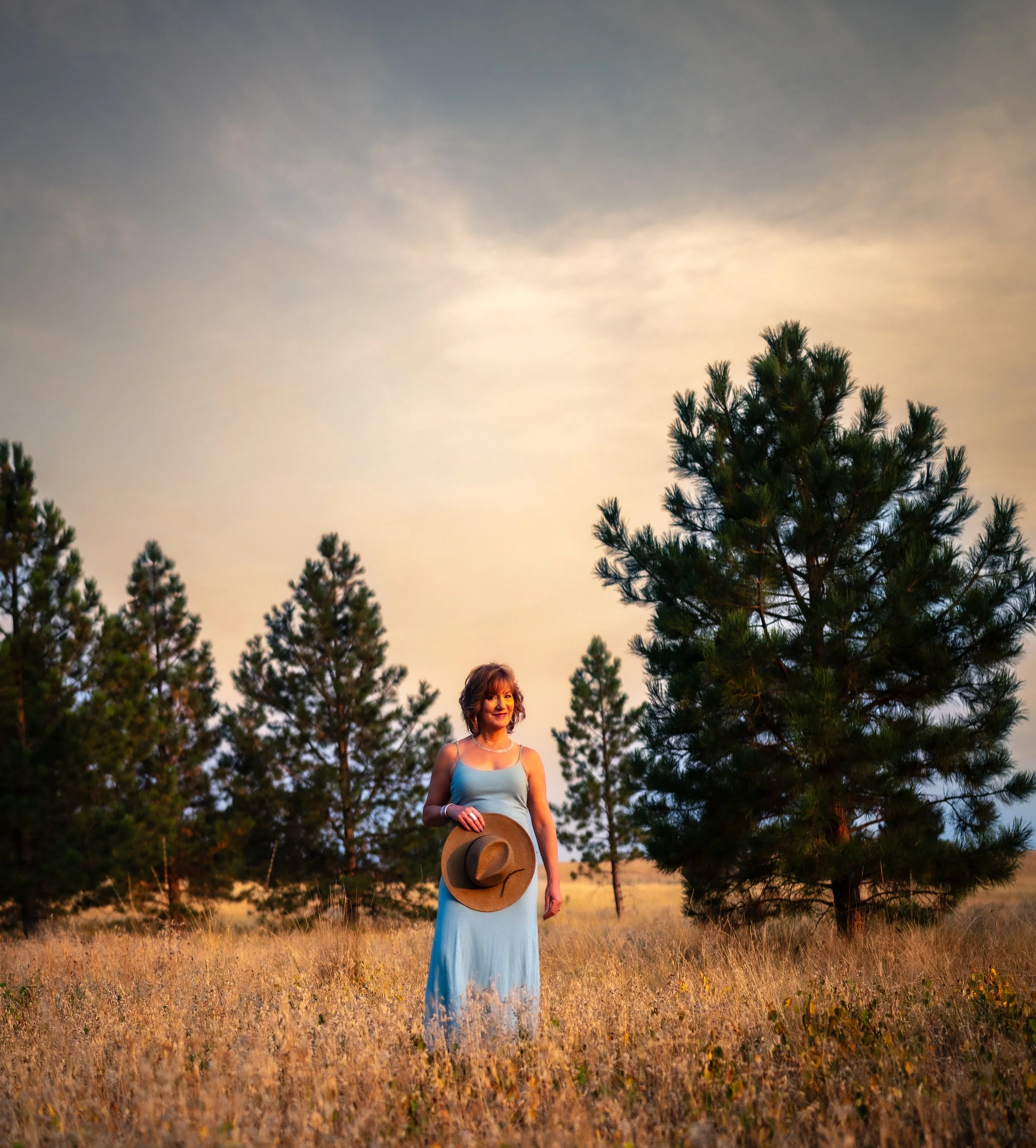 A woman standing in a field of dry grass, holding a wide-brimmed hat, with tall pine trees and a sunset sky in the background.